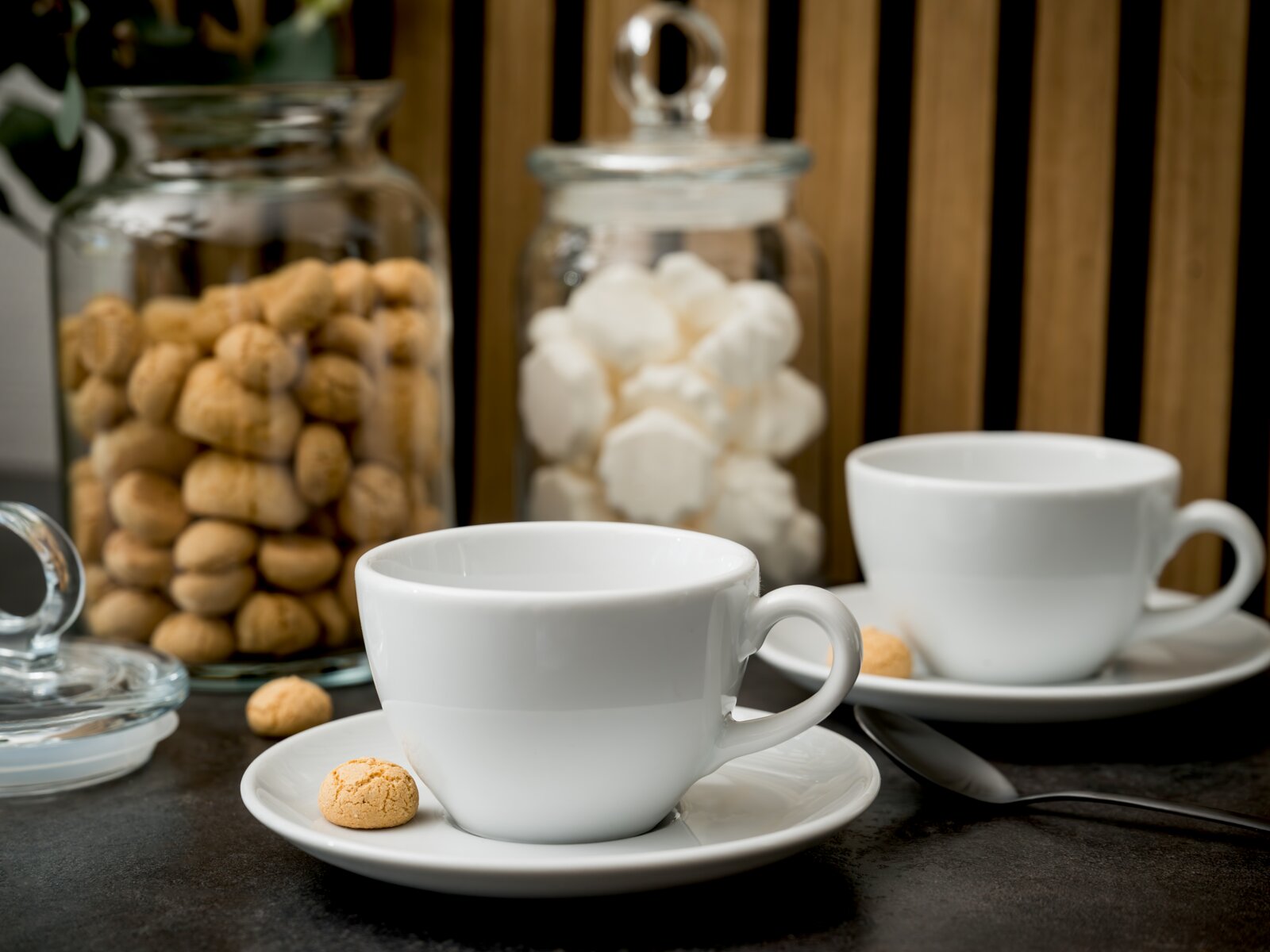 creaTable Cappuccino Set 8-tlg BISTROT Zwei weiße Porzellan-Kaffeetassen mit Untertassen auf dunkler Oberfläche, im Vordergrund eine Tasse mit kleinem Keks; im Hintergrund Glasbehälter mit Keksen und Zuckerwürfeln, Perspektive: leicht seitliche Nahaufnahme auf Tischhöhe