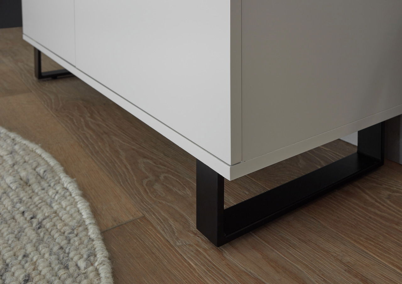 Nahaufnahme eines weißen Highboards mit schwarzen Metallbeinen auf einem Holzboden, seitliche Perspektive.