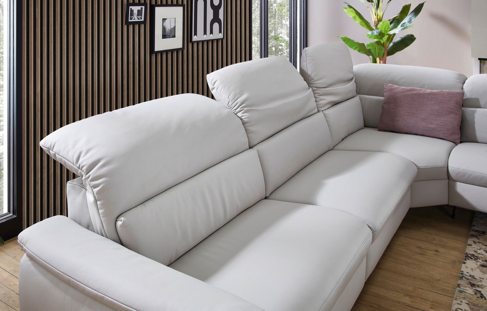 Eckgarnitur in hellem Leder mit verstellbaren Kopfstützen, seitlicher Perspektive, dekoriert mit einem lila Kissen, vor einer modernen Holzwand.