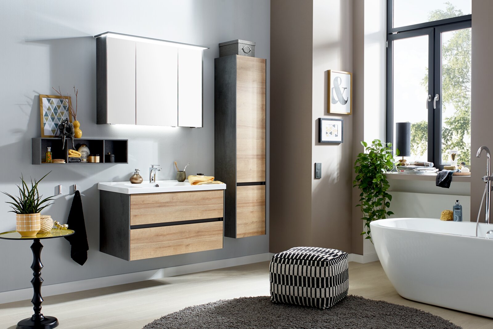 Modernes Badezimmer-Set mit wandhängendem Waschtischunterschrank in Holzoptik und Anthrazit, weißem Waschtisch, LED-Spiegelschrank und Hochschrank neben Badewanne am Fenster, Perspektive: frontale Raumansicht leicht von rechts.