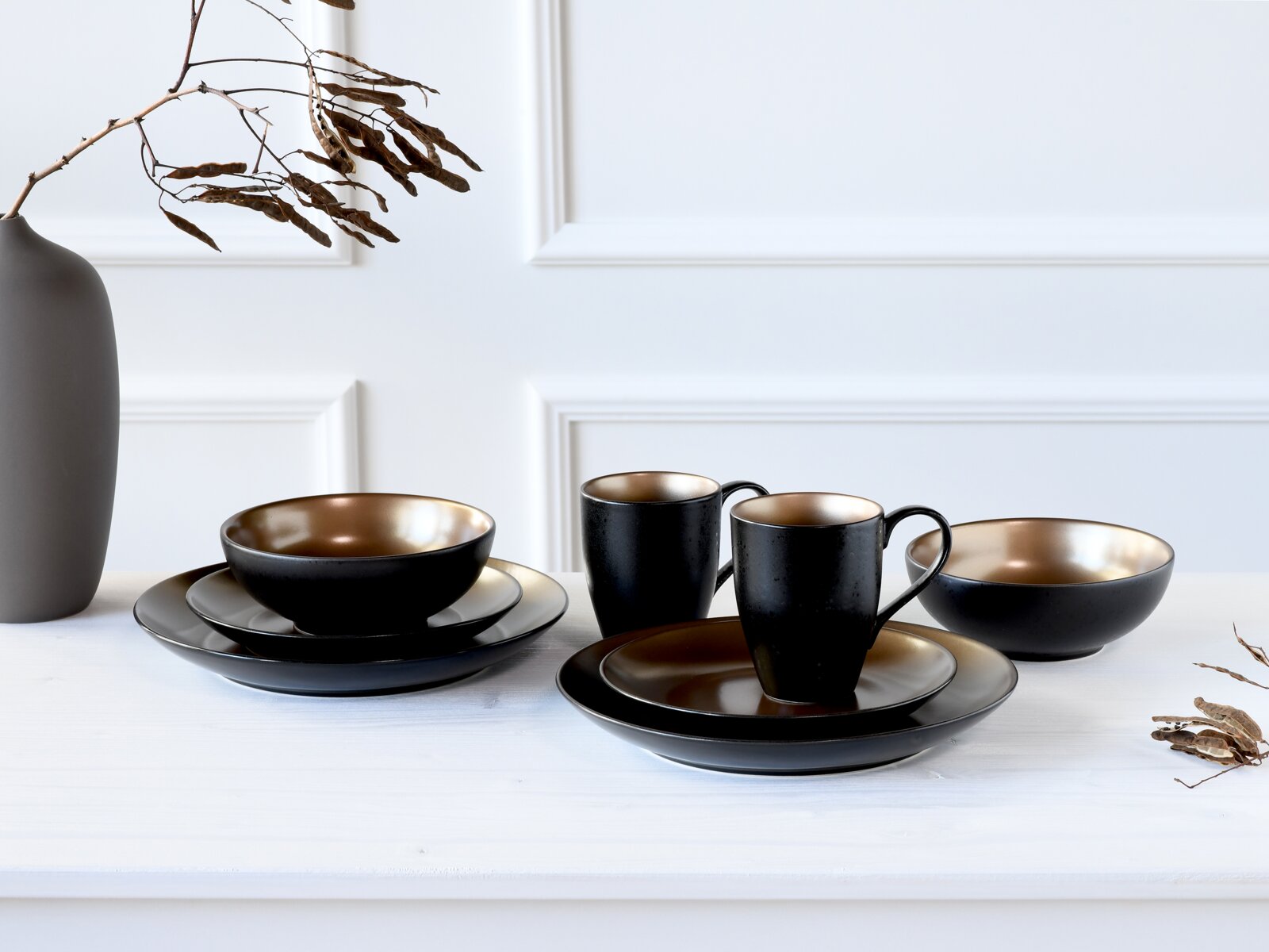 Schwarz-goldenes Keramik-Geschirrset mit zwei Tassen, Tellern und Schalen auf weißem Tisch vor heller Wand, perspektivisch in leichter Seitenansicht von vorn.