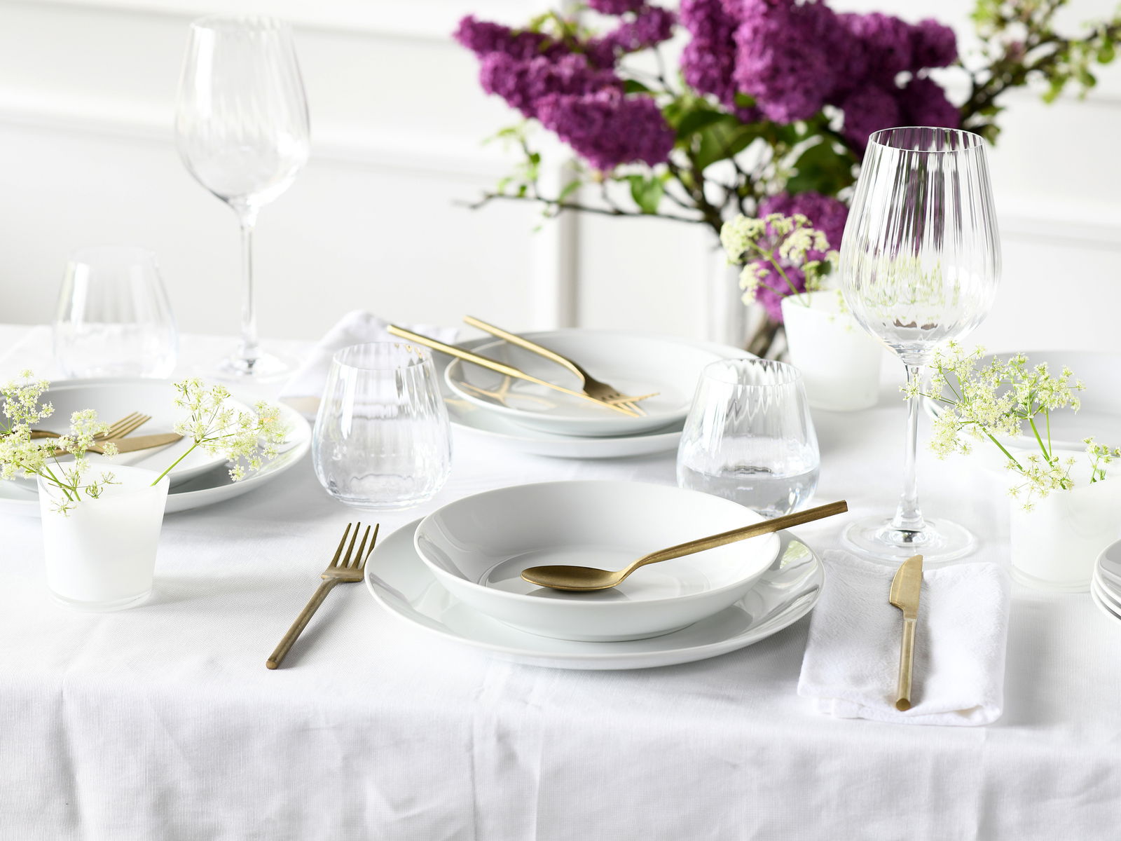 Elegantes Tellerset 18-tlg auf einem festlich gedeckten Tisch mit goldfarbenem Besteck und lila Blumen im Hintergrund, aus der Vogelperspektive fotografiert.