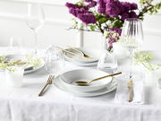 Elegantes Tellerset 18-tlg auf einem festlich gedeckten Tisch mit goldfarbenem Besteck und lila Blumen im Hintergrund, aus der Vogelperspektive fotografiert.