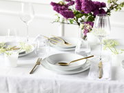 Elegantes Tellerset 18-tlg auf einem festlich gedeckten Tisch mit goldfarbenem Besteck und lila Blumen im Hintergrund, aus der Vogelperspektive fotografiert.