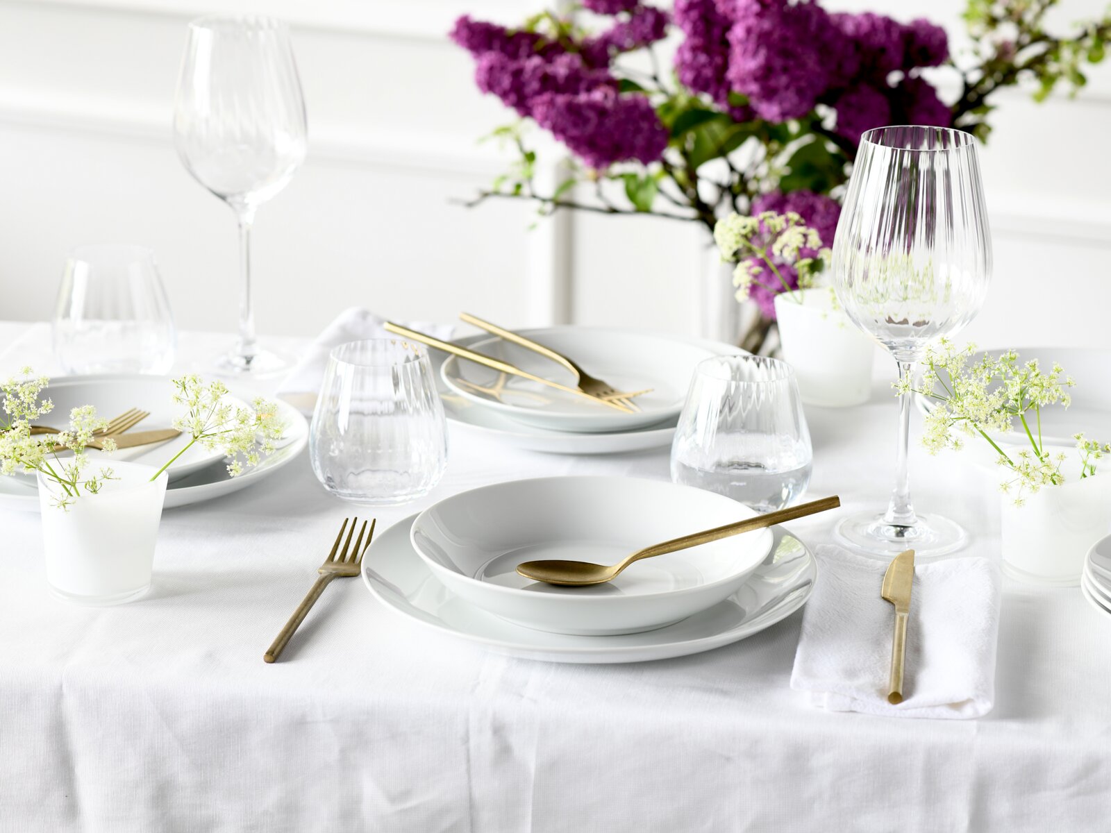Elegantes Tellerset 18-tlg auf einem festlich gedeckten Tisch mit goldfarbenem Besteck und lila Blumen im Hintergrund, aus der Vogelperspektive fotografiert.