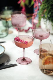 Rosa Sekt- und Dessertschale aus Glas, gefüllt mit einem Getränk und dekoriert mit rosa Blüten, auf einem Tisch mit weiteren Gläsern und Dekorationen, aus der Vogelperspektive fotografiert.