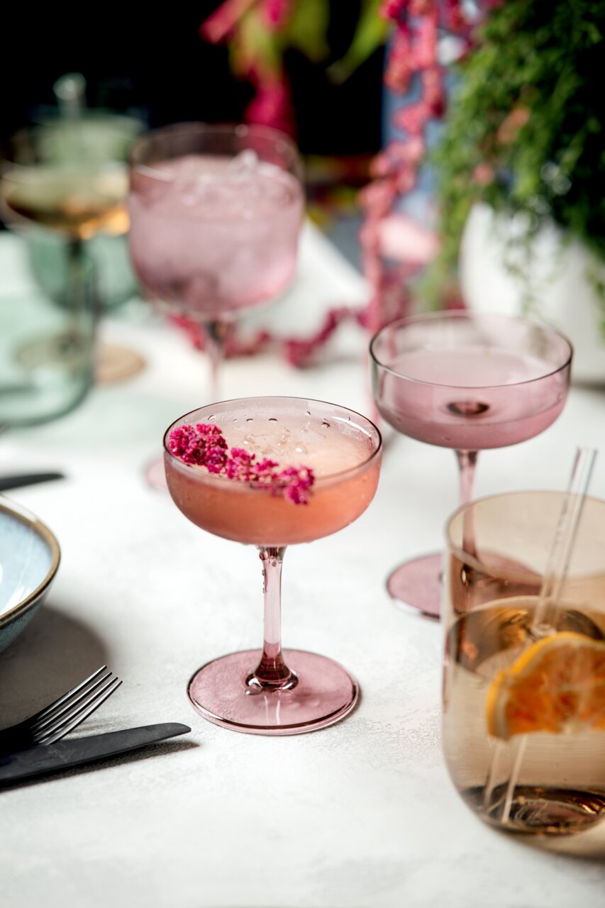Rosa Sekt- und Dessertschale aus Glas, gefüllt mit einem Getränk und dekoriert mit rosa Blüten, auf einem Tisch mit weiteren Gläsern und Dekorationen, aus der Vogelperspektive fotografiert.