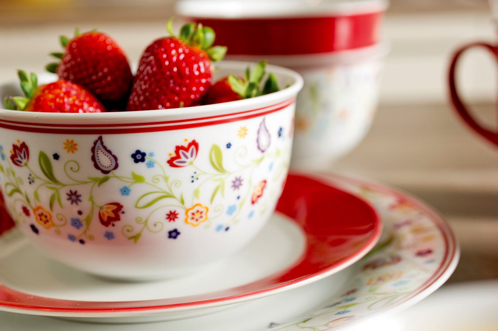 Nahaufnahme aus leicht erhöhter Frontalperspektive einer weißen Porzellanschale mit buntem Blumenmuster und rotem Rand, gefüllt mit frischen Erdbeeren und auf passendem Geschirr arrangiert