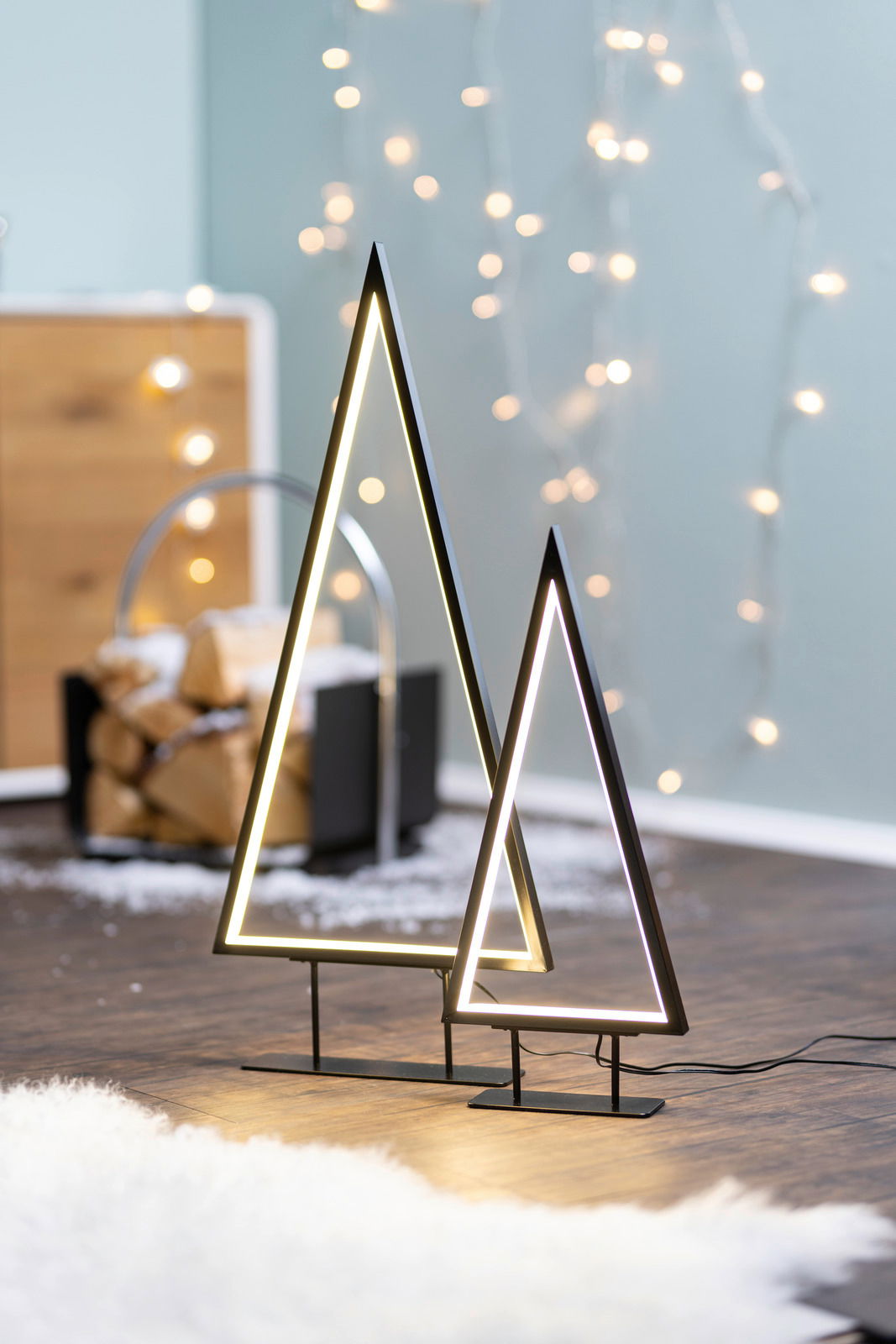 Zwei moderne LED-Bäume in Dreiecksform, stehend auf einem Holzboden, mit unscharfen Lichtern im Hintergrund. Seitliche Perspektive.