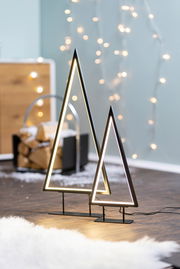 Zwei moderne LED-Bäume in Dreiecksform, stehend auf einem Holzboden, mit unscharfen Lichtern im Hintergrund. Seitliche Perspektive.