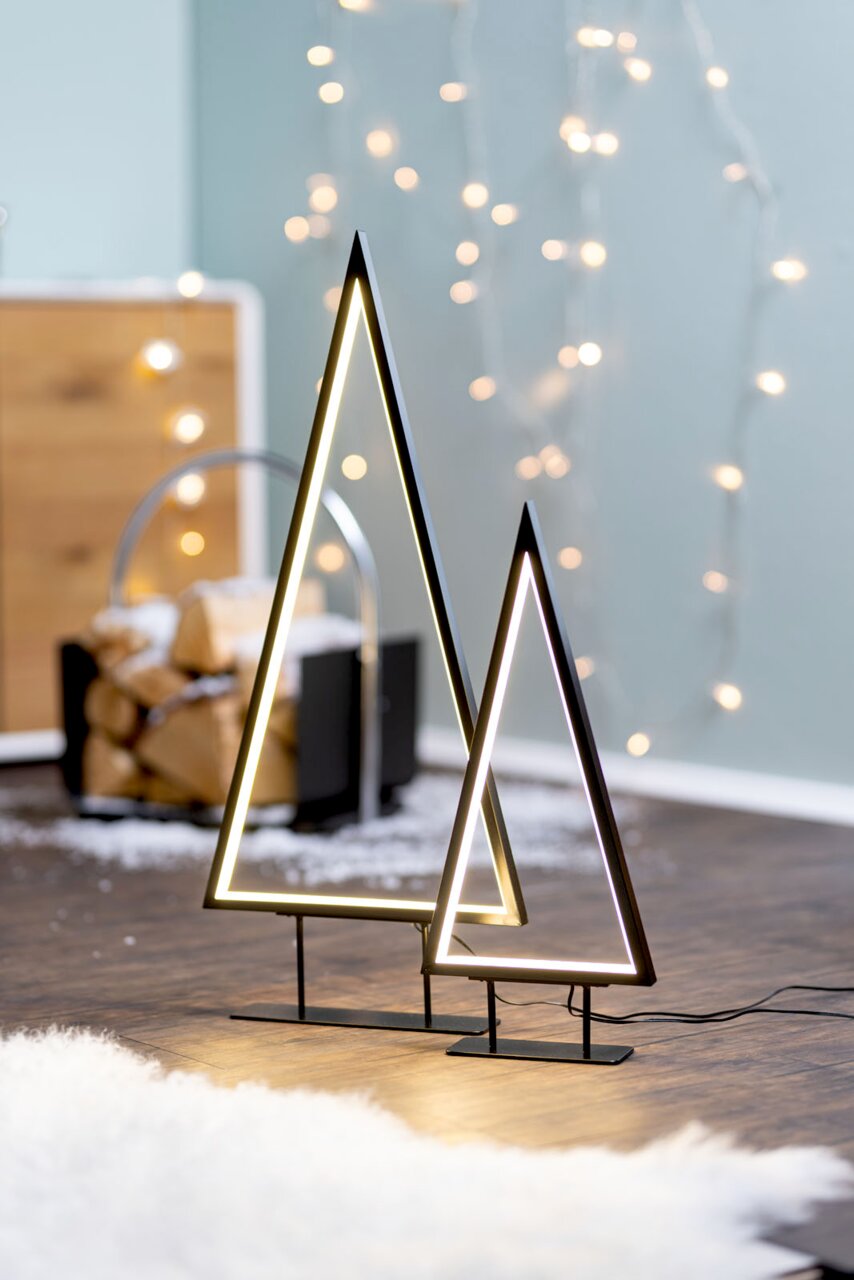 Zwei moderne LED-Bäume in Dreiecksform, stehend auf einem Holzboden, mit unscharfen Lichtern im Hintergrund. Seitliche Perspektive.