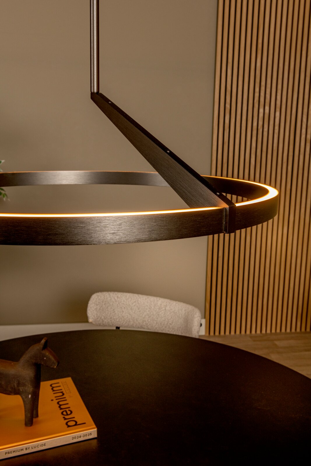 Moderne runde LED-Pendelleuchte in dunkler Metalloptik mit warmweiß leuchtendem Ring über einem Esstisch, perspektivisch in seitlicher Nahaufnahme von unten