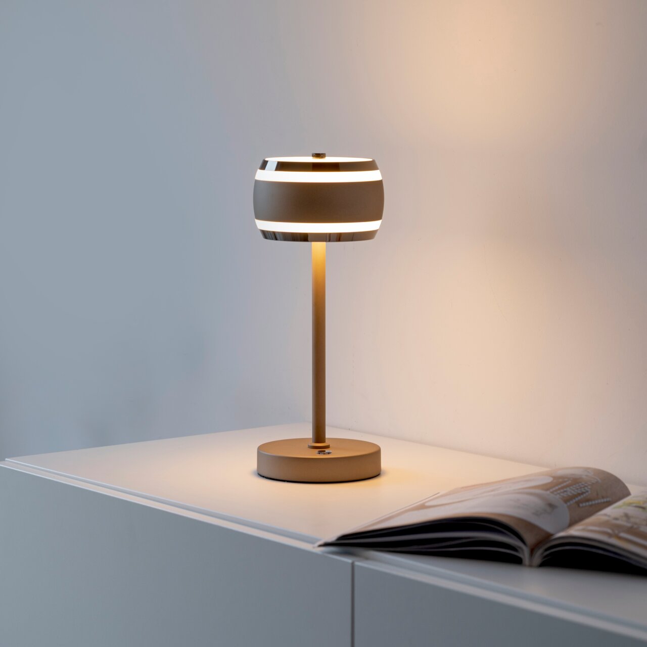 Moderne Akku-Tischleuchte in seitlicher Perspektive auf einem Tisch, mit einem eleganten, runden Lampenschirm und warmem Licht.