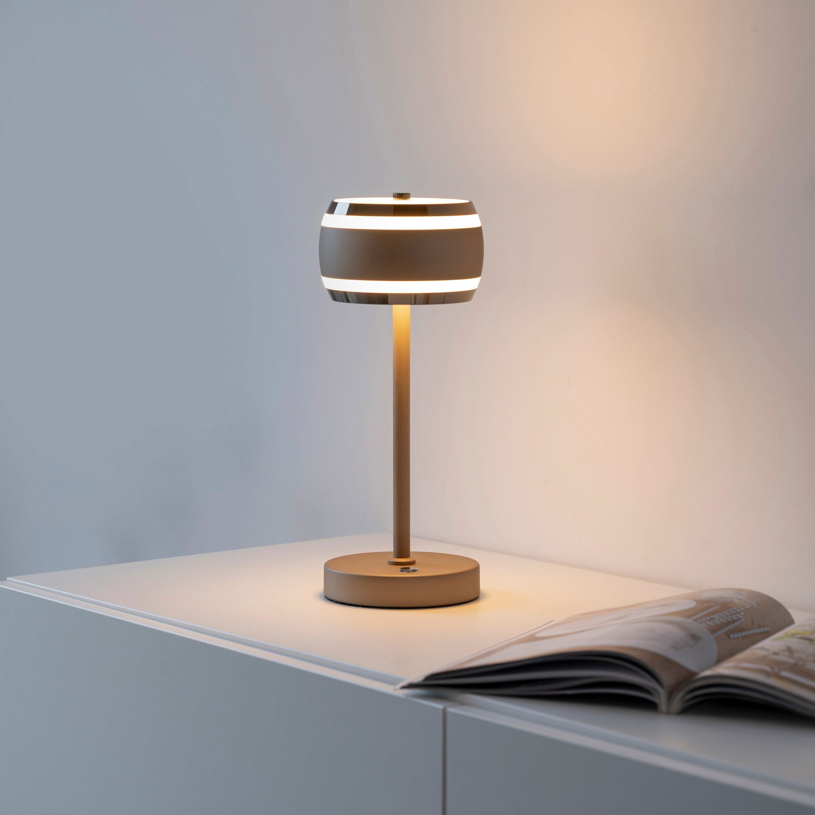 Moderne Akku-Tischleuchte in seitlicher Perspektive auf einem Tisch, mit einem eleganten, runden Lampenschirm und warmem Licht.