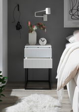 Moderne Nachtkommode mit zwei Schubladen in Weiß mit geriffelter Oberfläche, auf schwarzem Metallgestell. Perspektive: Frontalansicht.