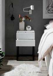 Moderne Nachtkommode mit zwei Schubladen in Weiß mit geriffelter Oberfläche, auf schwarzem Metallgestell. Perspektive: Frontalansicht.