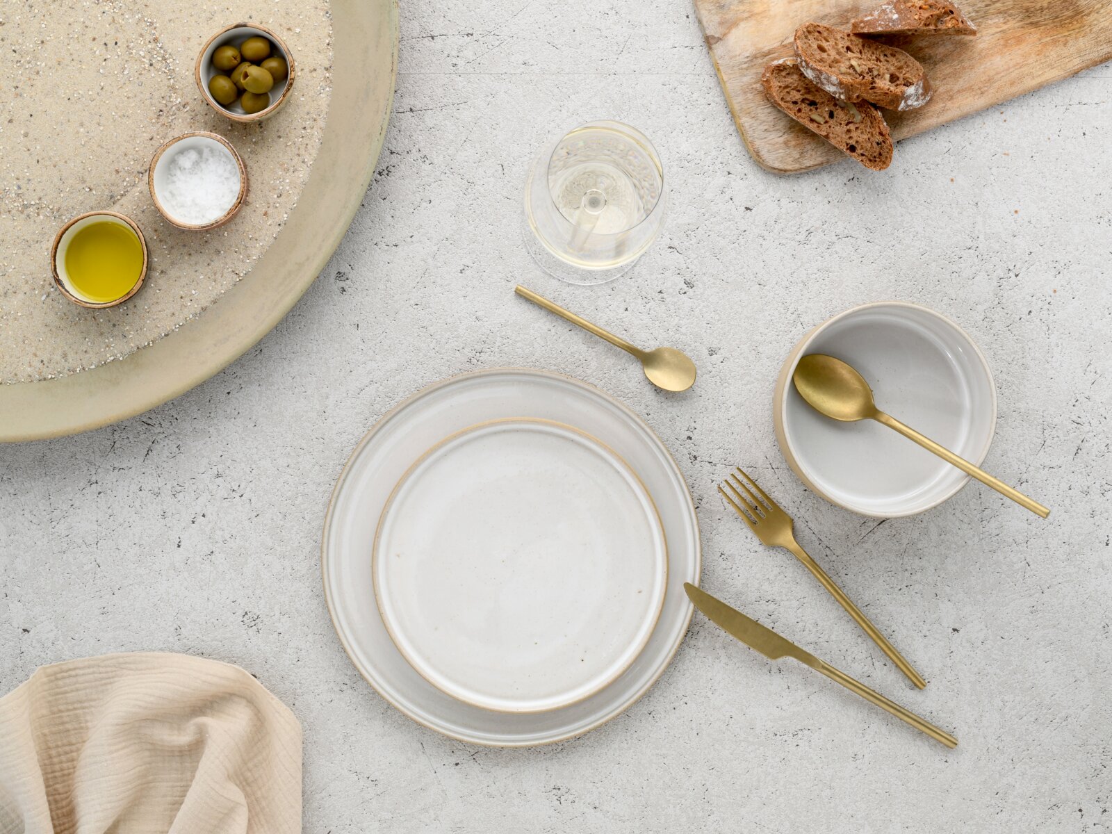 Draufsicht auf ein minimalistisches Tisch-Arrangement mit weißen Keramiktellern und -schale, goldfarbenem Besteck, Weinglas, Oliven, Salz, Olivenöl und Brotscheiben auf hellem Steinuntergrund