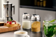 Zwei Zucker- oder Milchdosierer TIP aus Glas und Edelstahl, gefüllt mit Milch und braunem Zucker, auf einer Küchenarbeitsplatte. Perspektive von vorne.