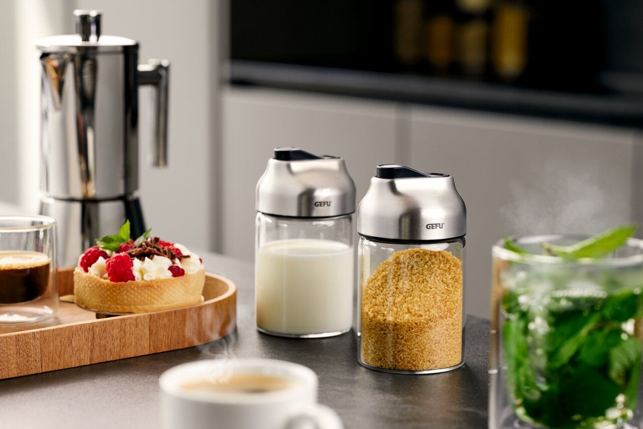 Zwei Zucker- oder Milchdosierer TIP aus Glas und Edelstahl, gefüllt mit Milch und braunem Zucker, auf einer Küchenarbeitsplatte. Perspektive von vorne.
