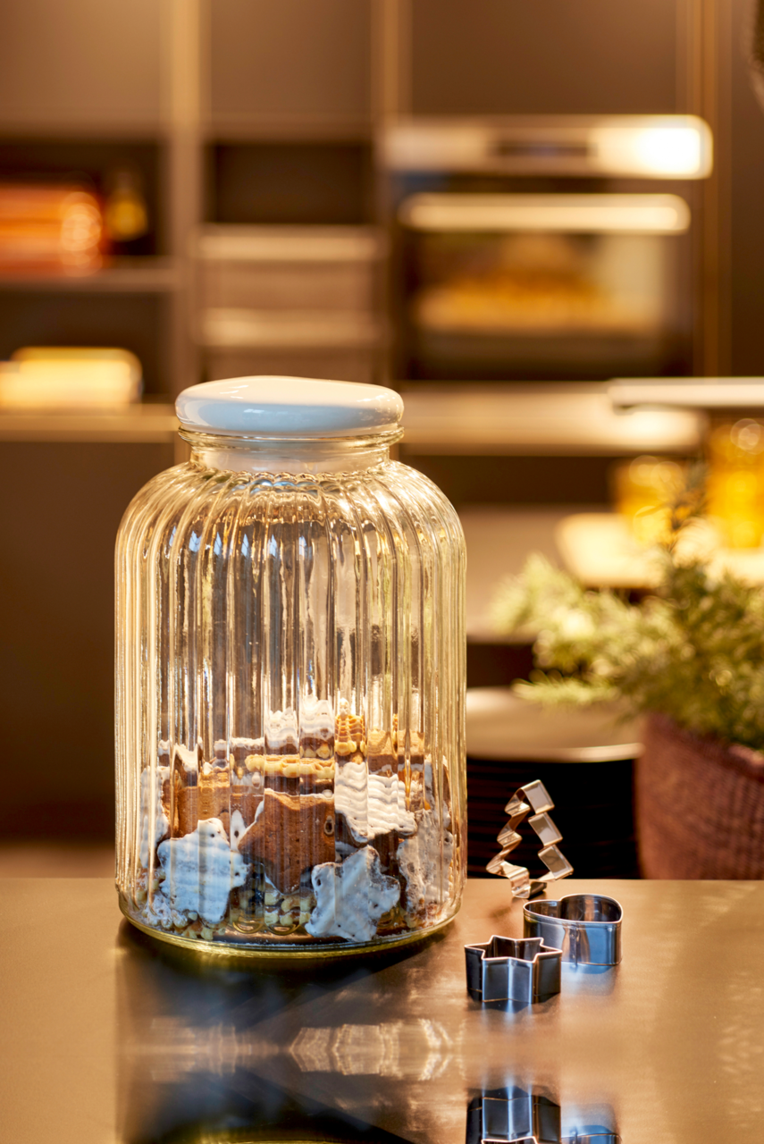 Großes, geriffeltes Glasgefäß mit weißem Deckel, gefüllt mit Keksen in Sternform, auf einem Küchentisch. Daneben liegen Ausstechformen in verschiedenen Formen. Perspektive: seitlich.