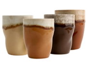 Vier glasierte Keramikbecher in Naturtönen (Beige, Braun, Dunkelbraun, Terrakotta) mit zweifarbiger Glasur auf weißem Hintergrund, frontal in leichter Schrägansicht fotografiert.
