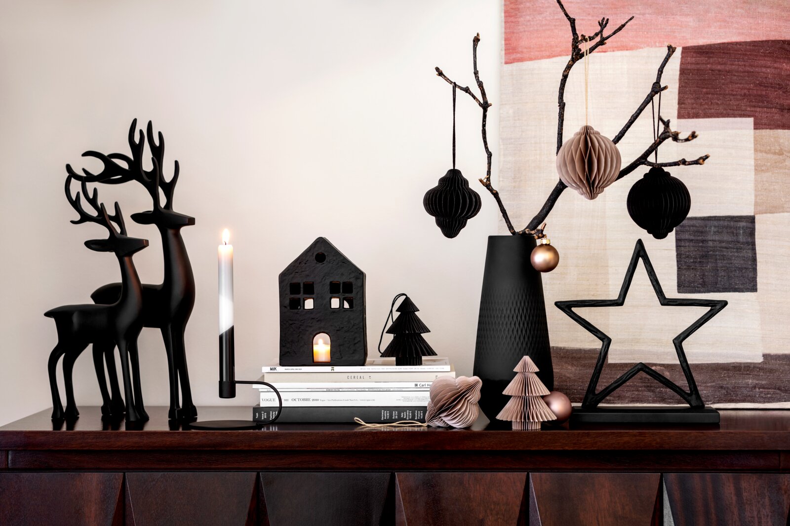 Frontalansicht einer festlich dekorierten Holzkommode mit schwarzen Hirschfiguren, Kerzen, Papierornamenten, Zweigen in einer Vase und großem Metallstern in elegantem Schwarz und Rosé.