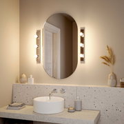 Badezimmer mit ovalem Spiegel, flankiert von zwei LED-Leuchtmitteln, die warmes Licht ausstrahlen. Perspektive: Frontalansicht auf den Spiegel und das Waschbecken.