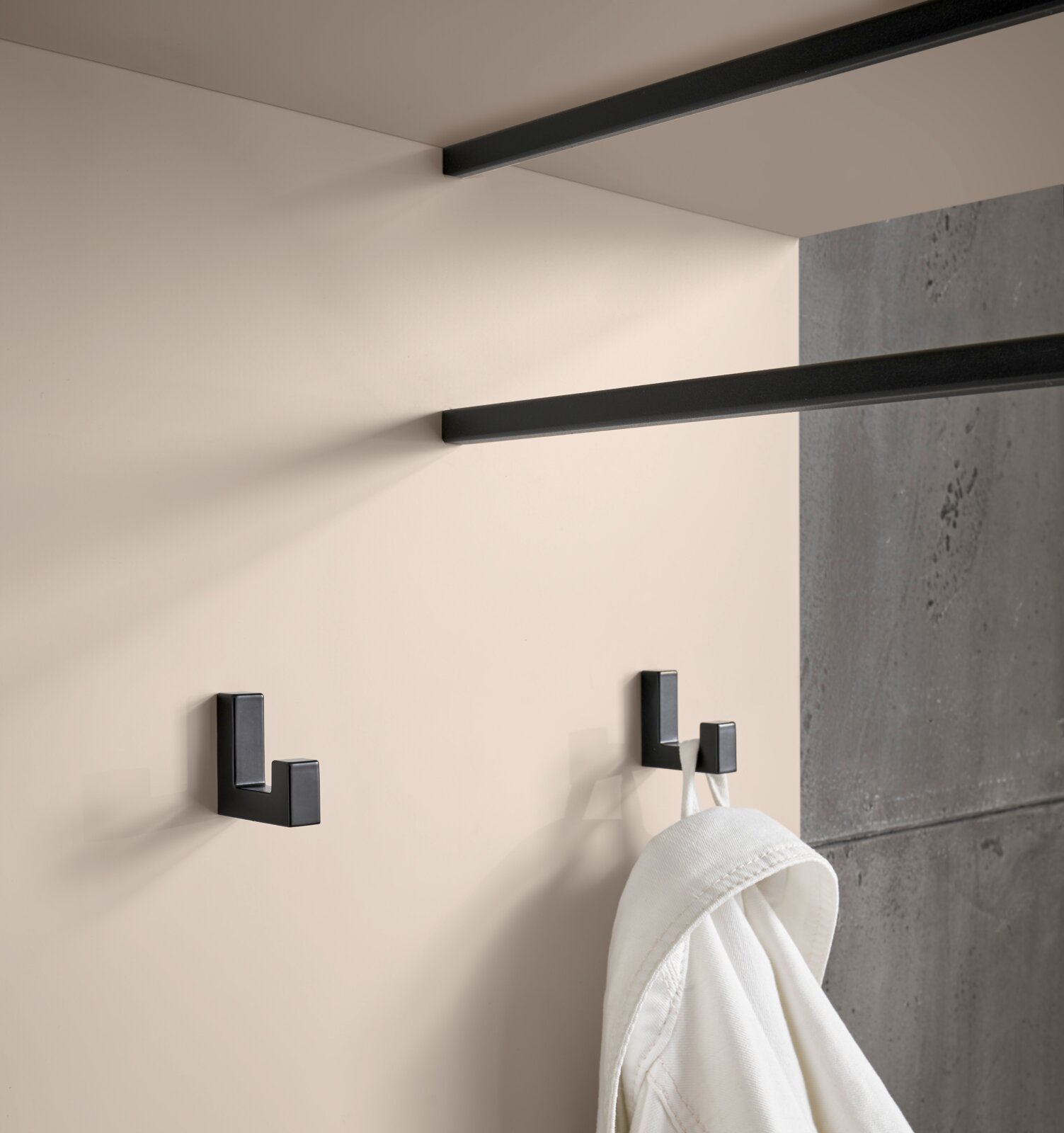 Schwarzer, eckiger Wandhaken im modernen Design an heller Wand, seitliche Nahaufnahme in leicht schräger Perspektive; daneben ein weiterer Haken mit aufgehängtem weißen Handtuch, Hintergrund in Betonoptik