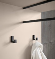 Schwarzer, eckiger Wandhaken im modernen Design an heller Wand, seitliche Nahaufnahme in leicht schräger Perspektive; daneben ein weiterer Haken mit aufgehängtem weißen Handtuch, Hintergrund in Betonoptik
