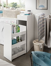 Waschmaschinenumbau-Set in einem modernen Waschraum, mit einem weißen, schmalen Regal auf Rollen, das neben einer Waschmaschine steht. Perspektive von vorne links.