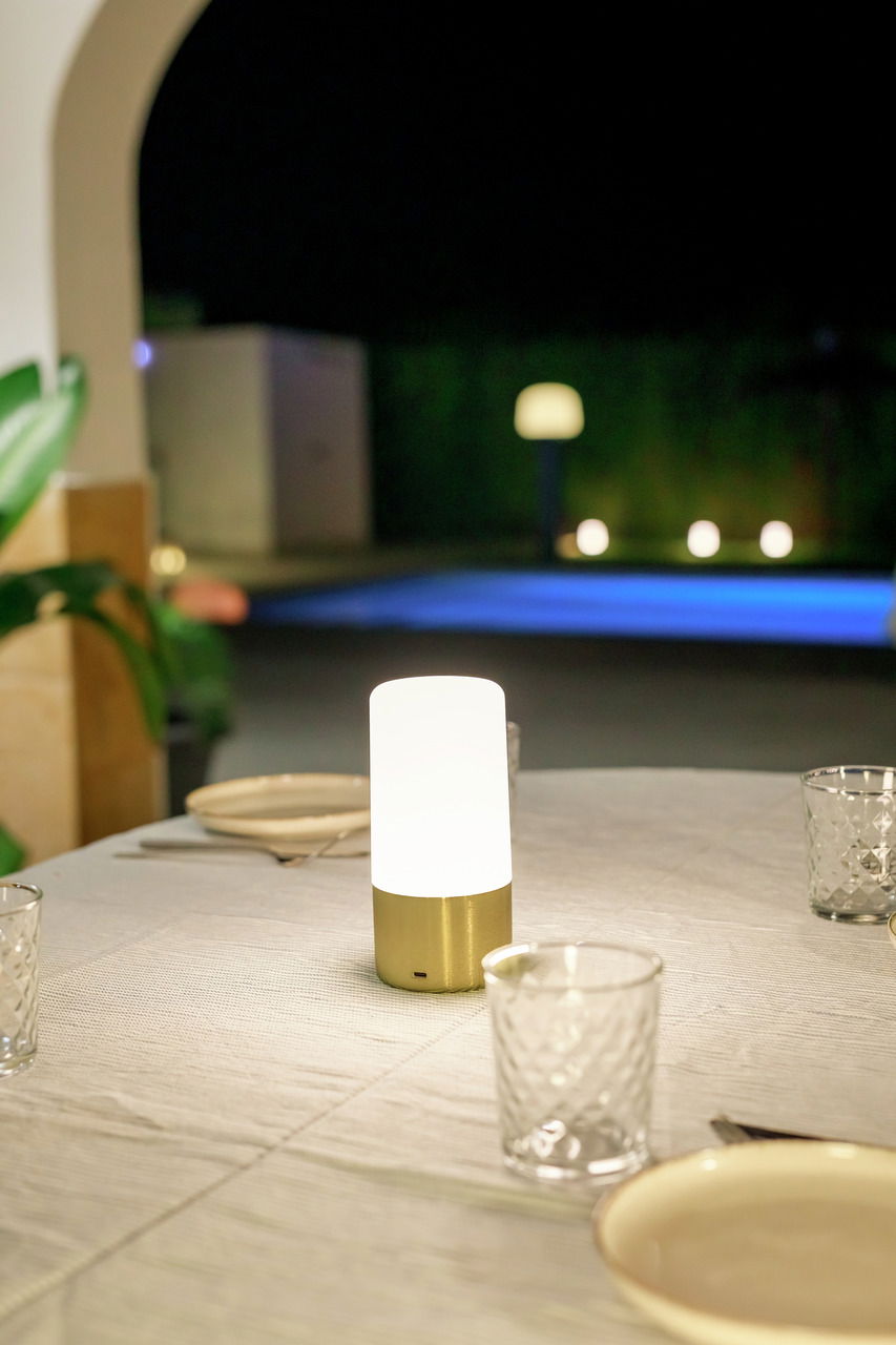 New Garden Akku-Tischleuchte MAIA Akku-Tischleuchte MAIA auf einem gedeckten Tisch im Freien, mit unscharfem Hintergrund und Poolbeleuchtung, seitliche Perspektive