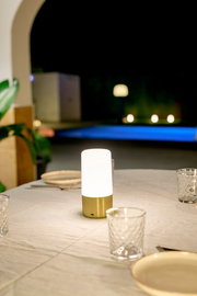 Akku-Tischleuchte MAIA auf einem gedeckten Tisch im Freien, mit unscharfem Hintergrund und Poolbeleuchtung, seitliche Perspektive