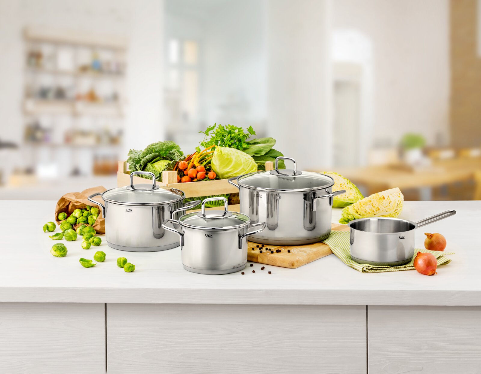 Silit 6-delige pannenset EXTRA COOK Set roestvrijstalen kookpannen, bestaande uit vier pannen met deksels, op een aanrecht met verse groenten op de achtergrond, gezien vanuit een vooraanzicht.