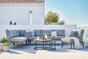 Moderne Ecklounge mit grauen Kissen und zwei runden Loungetischen auf einer Terrasse, seitliche Perspektive.