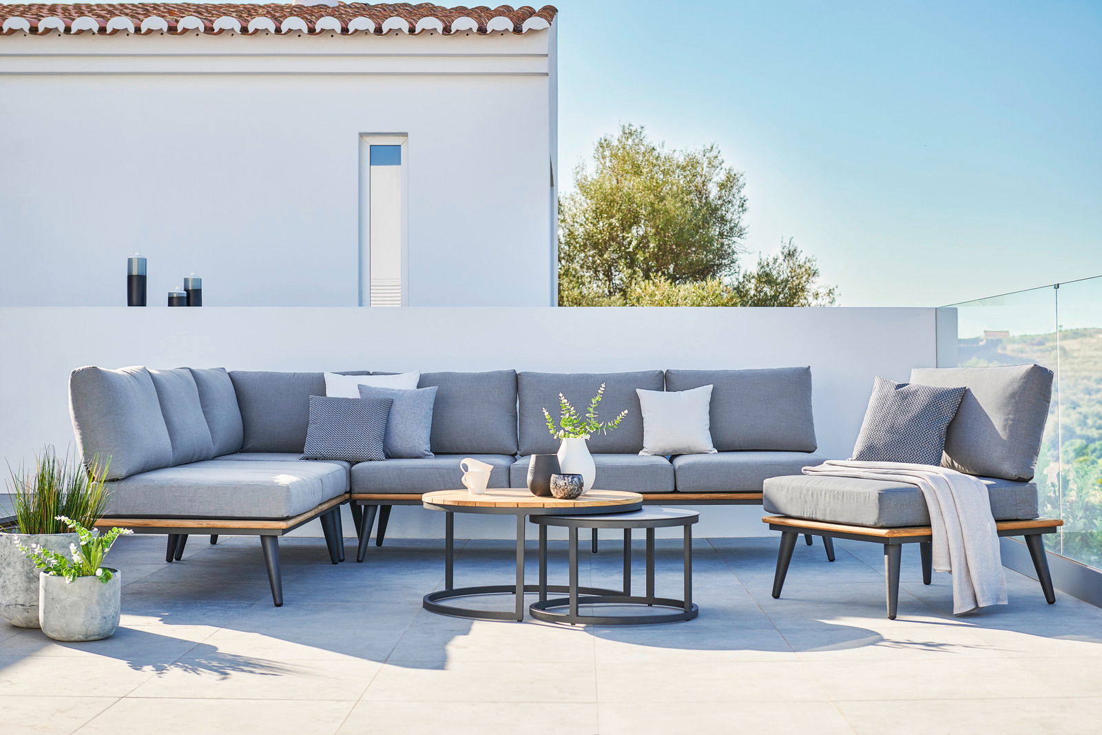 Moderne Ecklounge mit grauen Kissen und zwei runden Loungetischen auf einer Terrasse, seitliche Perspektive.