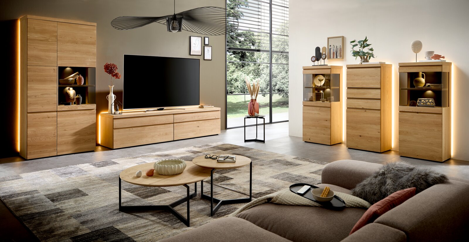 Modernes Wohnzimmermöbel-Set aus hellem Massivholz mit TV-Lowboard, Vitrinen, Highboard und beleuchteten Schränken, präsentiert in frontaler Weitwinkelansicht in einem stilvollen Wohnambiente.