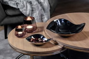 Zwei runde Holztische mit einem schwarzen, geschwungenen Teelichthalter aus Glas in Sandfarbe, der von oben betrachtet wird. Daneben stehen zwei weitere Teelichthalter mit brennenden Kerzen.