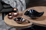 Zwei runde Holztische mit einem schwarzen, geschwungenen Teelichthalter aus Glas in Sandfarbe, der von oben betrachtet wird. Daneben stehen zwei weitere Teelichthalter mit brennenden Kerzen.