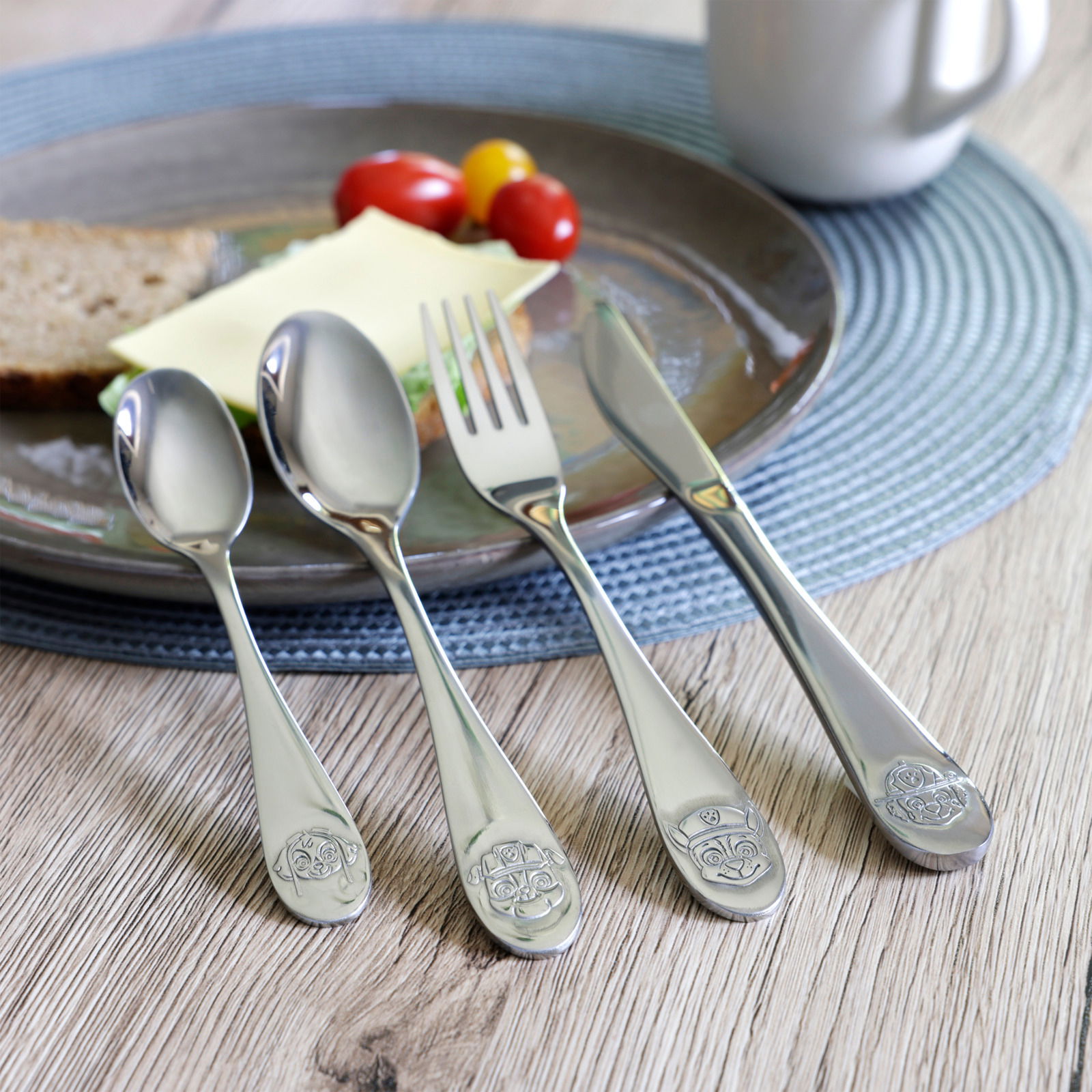 4-teiliges Edelstahlbesteckset mit kindgerechten Motiven, bestehend aus Löffel, Gabel und Messer, auf einem Holztisch neben einem gedeckten Teller, aus der Vogelperspektive fotografiert.
