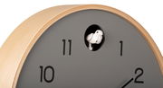 Nahaufnahme einer modernen Wanduhr mit grauem Zifferblatt und hellem Holzrahmen. Ein kleiner weißer Kuckucksvogel ist in einem runden Fenster über der Zahl 12 sichtbar.