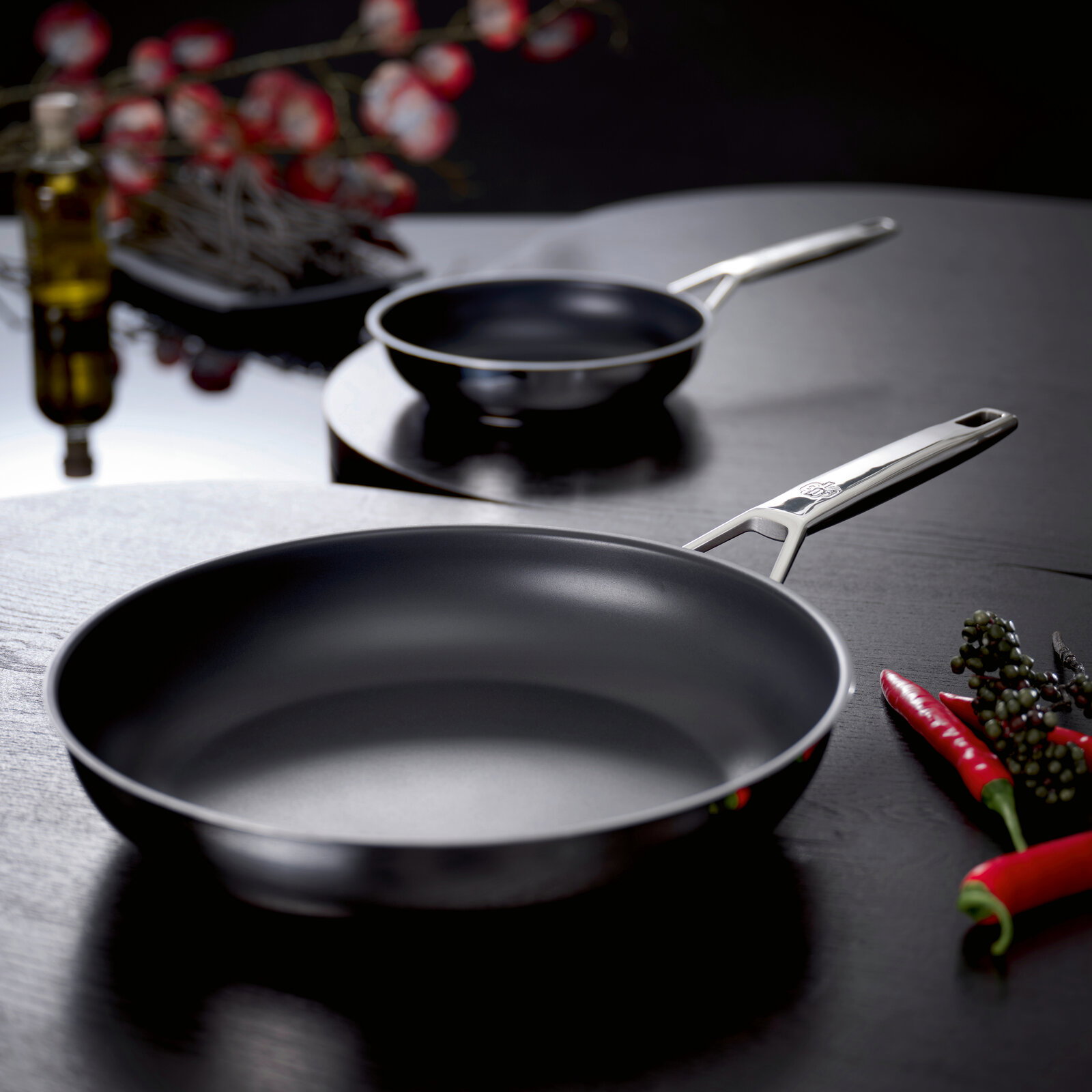 Schulte-Ufer Bratpfanne ASTRAL Zwei schwarze Pfannen mit Edelstahlgriffen auf dunklem Tisch, vorn eine große Bratpfanne und hinten eine kleinere Stielkasserolle, fotografiert aus leicht erhöhter schräger Perspektive