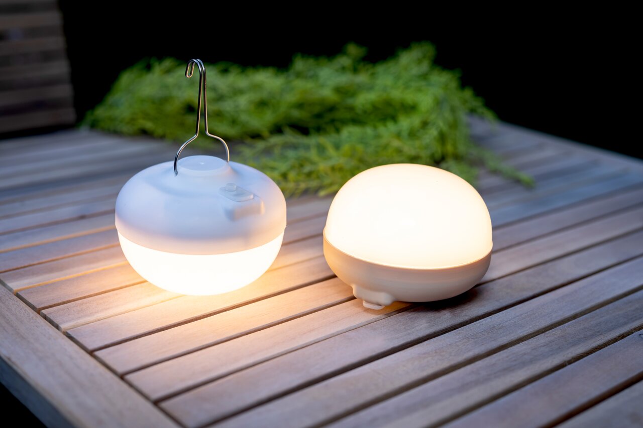 Twee ronde CHERRY-lampen op accu van New Garden op een houten tafel, waarvan één met een haak om op te hangen, gefotografeerd vanuit een zijaanzicht.