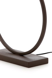 Close-up van een moderne tafellamp met een ovale ring en een rechthoekige voet in donkerbruin, zijaanzicht van de voet met zichtbare kabel tegen een witte achtergrond
