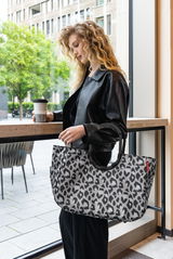 Frau mit lockigem Haar hält eine große Tasche mit Leopardenmuster in der Hand, während sie an einem Fenster steht. Die Tasche ist in einer seitlichen Perspektive zu sehen.