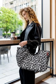 Frau mit lockigem Haar hält eine große Tasche mit Leopardenmuster in der Hand, während sie an einem Fenster steht. Die Tasche ist in einer seitlichen Perspektive zu sehen.