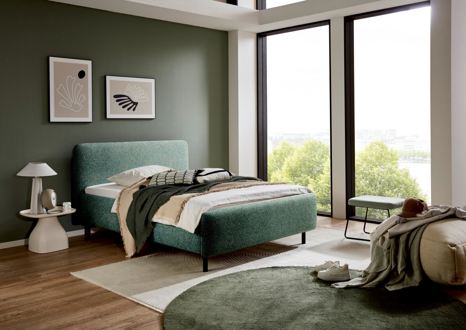 Modernes Polsterbett in einem stilvollen Schlafzimmer mit grüner Wand und großen Fenstern, seitliche Perspektive.