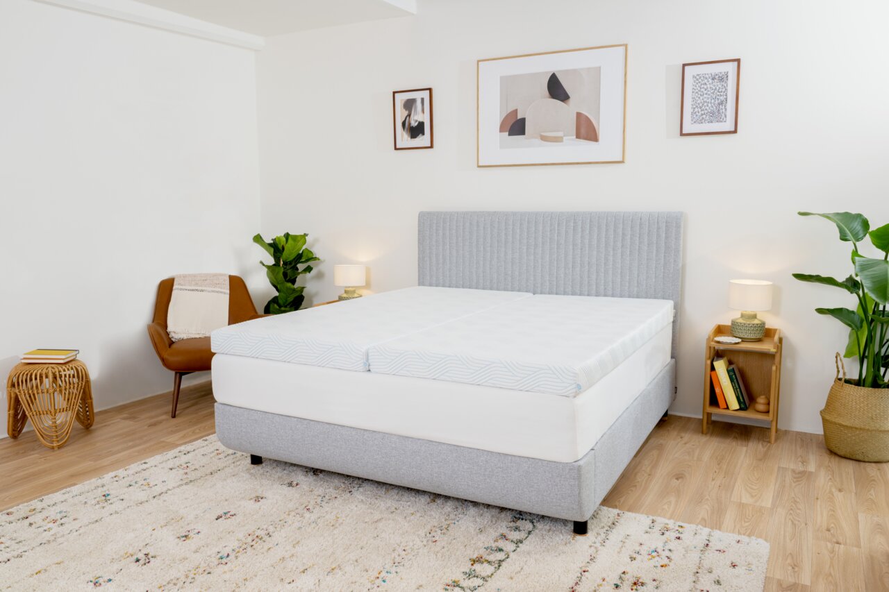 Schlafzimmer mit einem grauen Boxspringbett, das mit einem Topper Firm ausgestattet ist. Das Bett steht in einem modernen Raum mit Pflanzen, einem Sessel und Wanddekorationen. Perspektive von der Seite des Bettes.