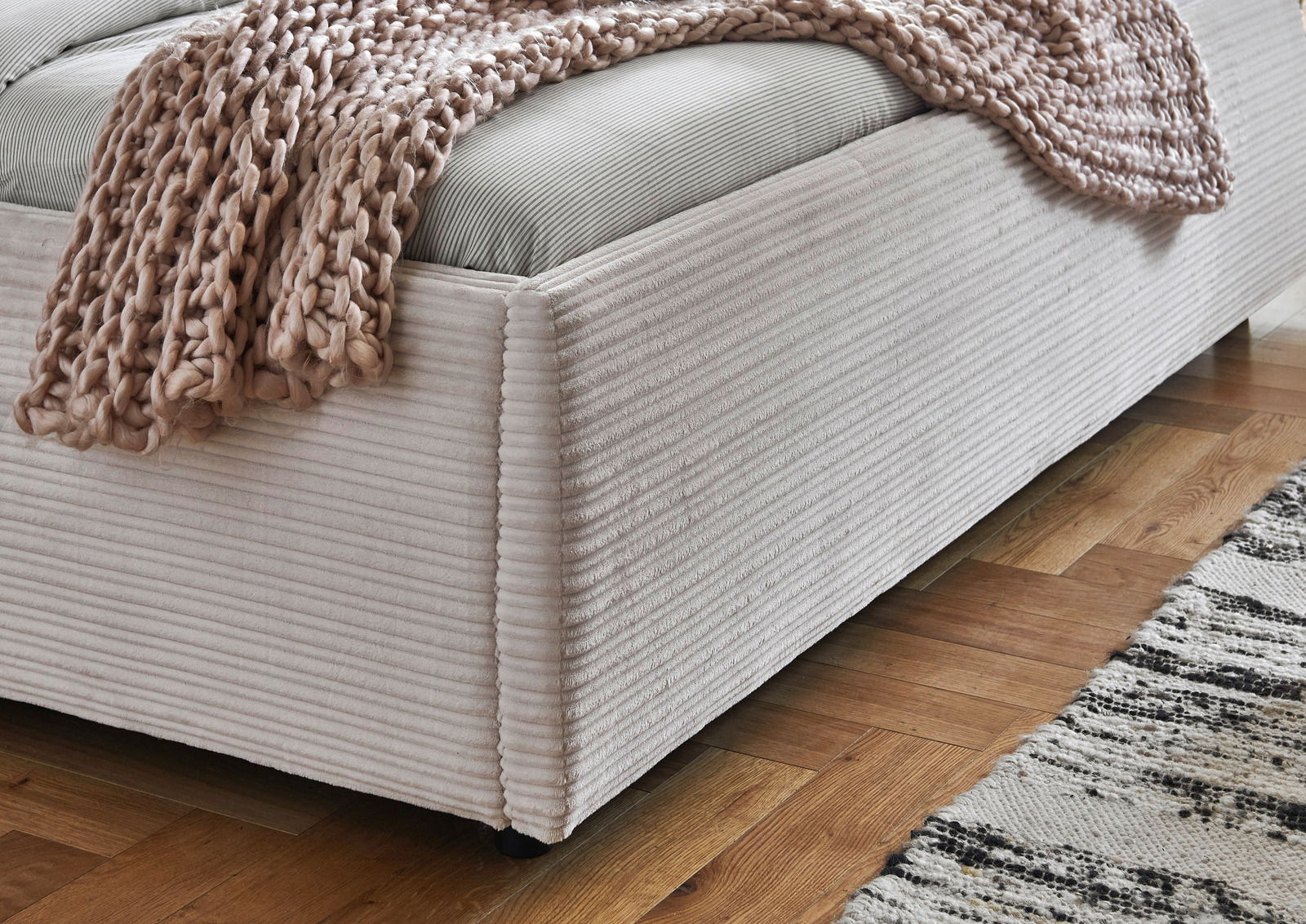 Nahaufnahme eines Polsterbetts mit geripptem Stoffbezug und einer grob gestrickten Decke, seitliche Perspektive