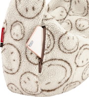 Nahaufnahme in schräger Vorderansicht: flauschiger Reißverschluss-Rucksack aus cremefarbenem Teddy-Material mit braunem Smileymuster, Fronttasche mit Reißverschluss und teilweise sichtbarem Smartphone