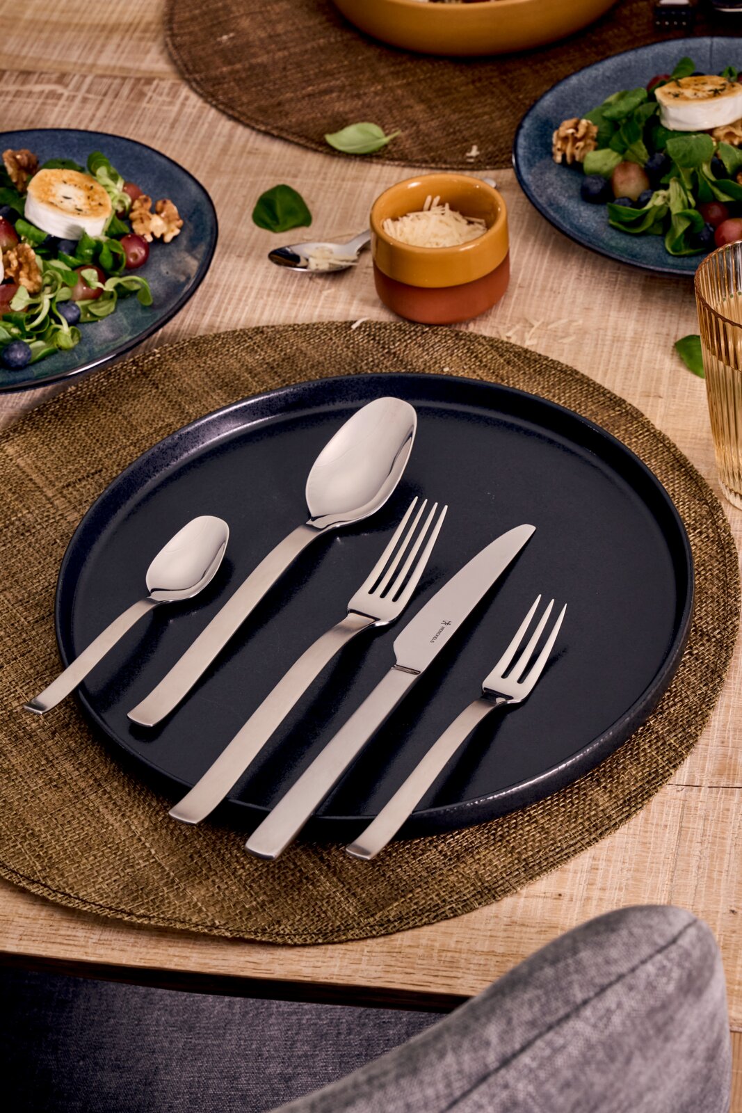 Roestvrijstalen bestekset met twee vorken, mes en twee lepels op een donkerblauw bord op een gedekte eettafel, gefotografeerd in schuin bovenaanzicht.